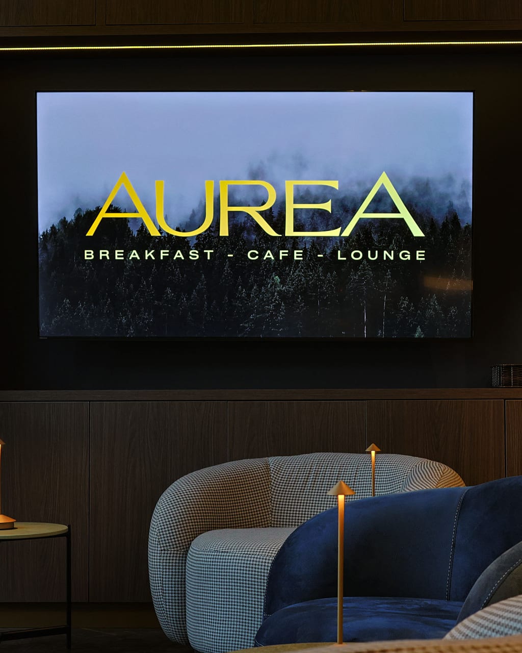 Die Lounge der Auréa: der Auréa-Bildschirm über Samt- und Hahnentritt-Sesseln mit goldenen Tischleuchten